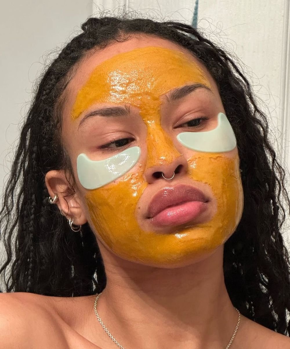 Rosto de pessoa com máscara facial de argila amarela e adesivos sob os olhos.