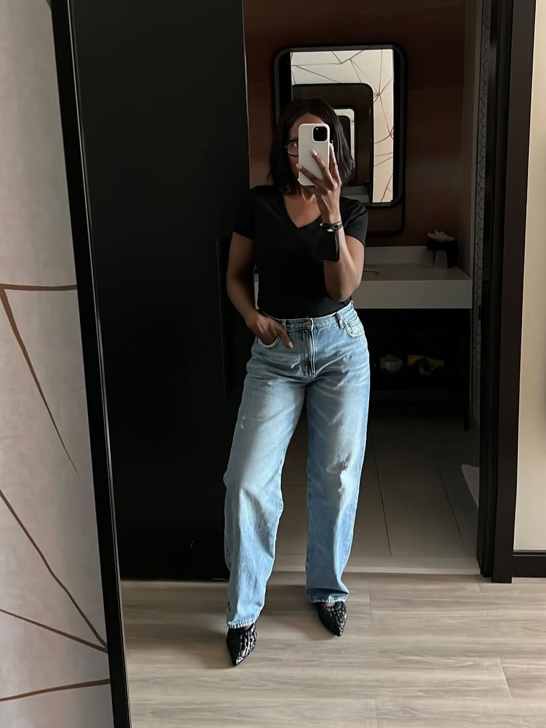 Pessoa de blusa gola V, calça jeans, posando em frente ao espelho com celular na mão.