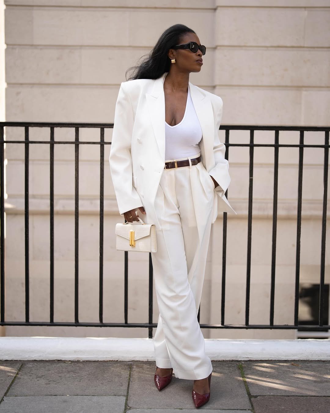 Mulher elegante com terno branco e blusa gola V, segurando bolsa e usando óculos escuros.
