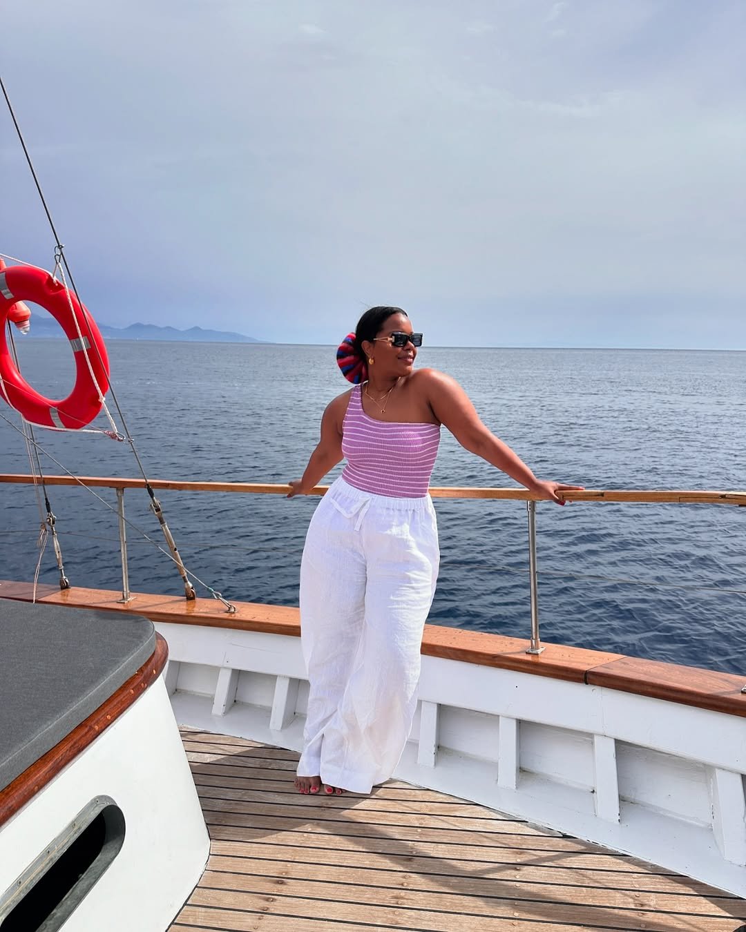 Pessoa com blusa listrada roxa e calça branca apoia-se na grade de um barco, com o mar ao fundo.