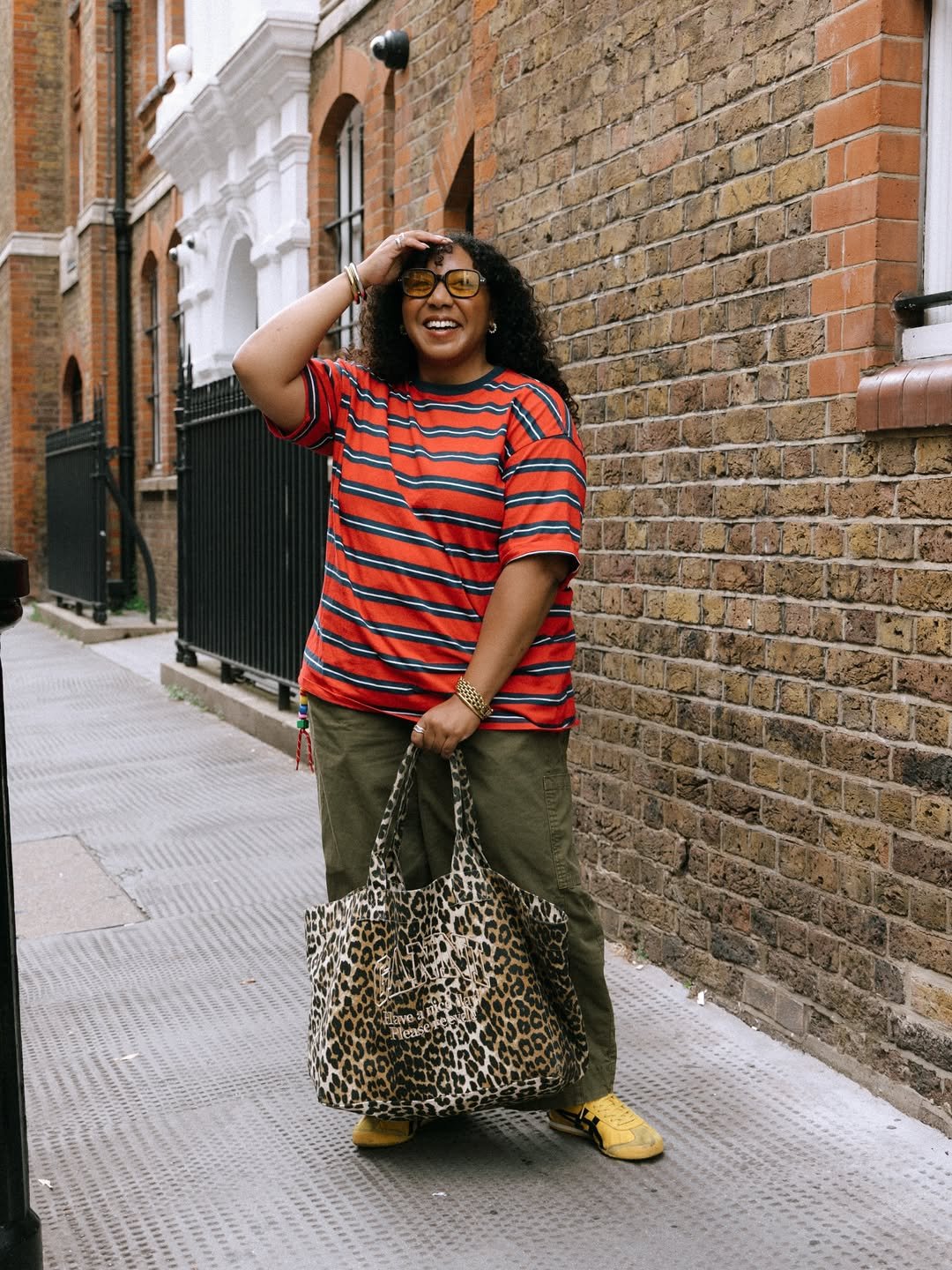 Pessoa sorridente usando blusa listrada vermelha e segurando bolsa estampada, em uma rua de tijolos.