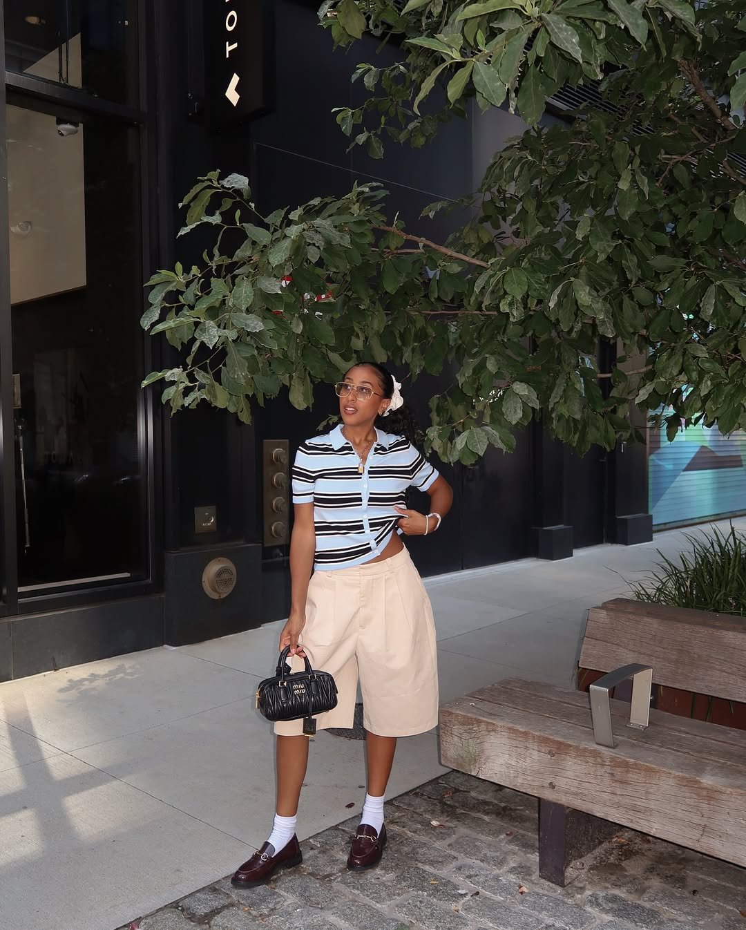 Pessoa usando blusa listrada e bermuda bege, segurando uma bolsa preta, em calçada ao lado de árvore.