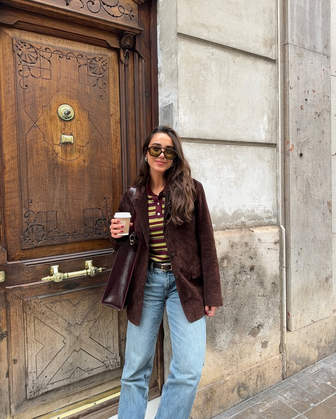 Pessoa com blusa listrada marrom e amarela, jaqueta marrom e óculos escuros, segurando café em frente a porta de madeira ornamentada.