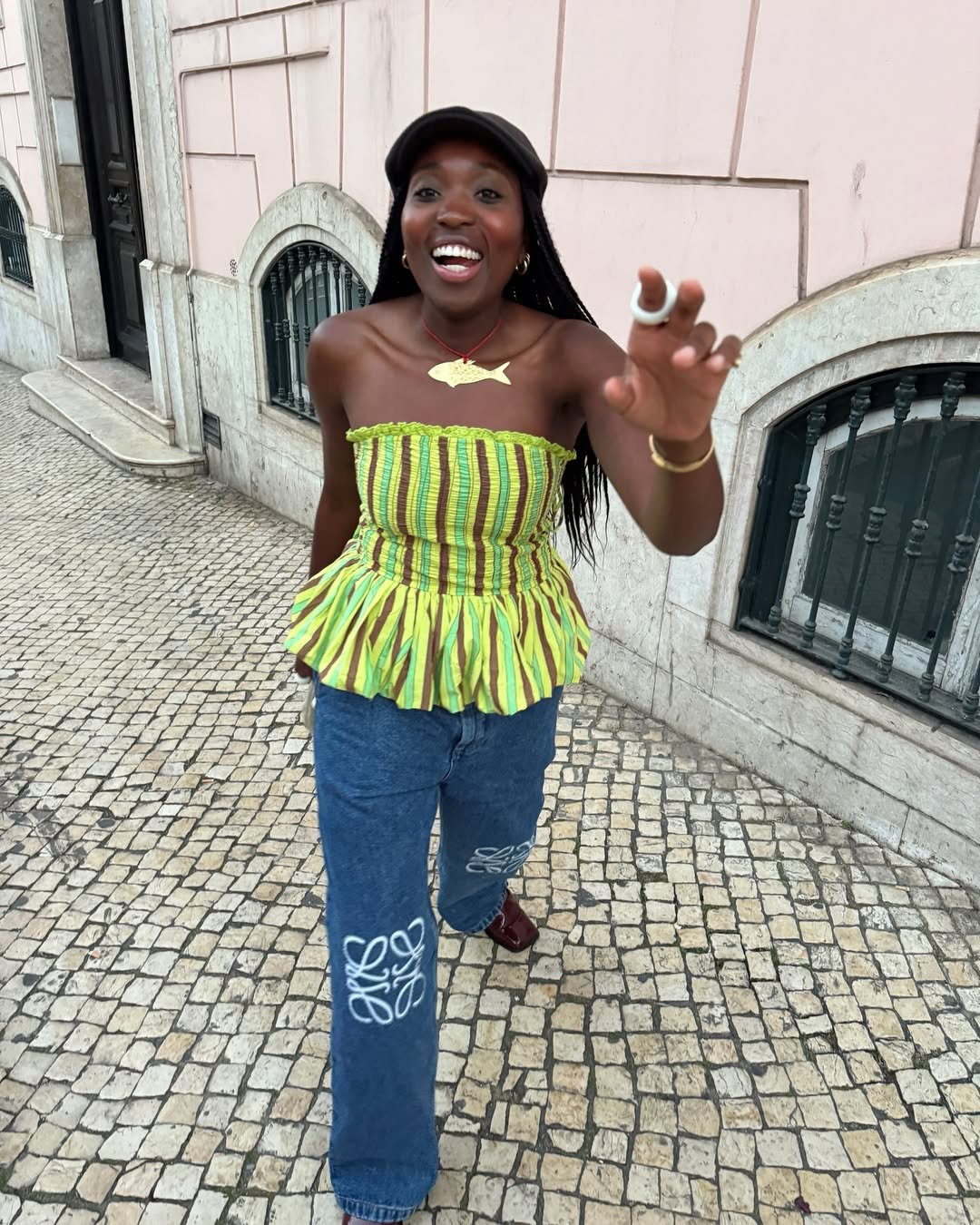 Pessoa sorridente usando blusa listrada verde e amarela na calçada de pedra, com jeans solto e boné.