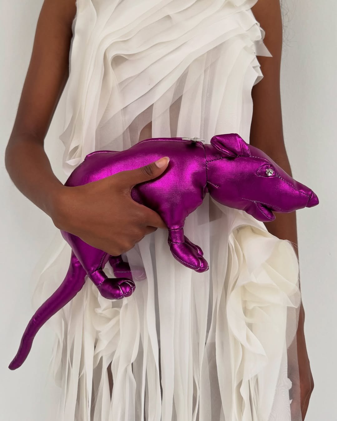 Pessoa segurando uma bolsa de rato roxa metálica, vestindo um vestido branco com babados.
