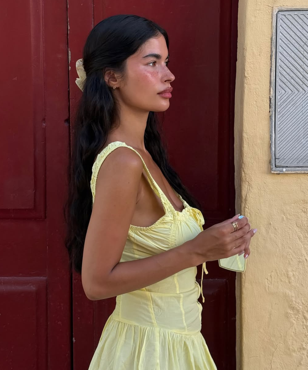 Mulher de perfil veste vestido amarelo claro, em frente a uma porta vermelha, segurando um celular. Pele lisa como manteiga.
