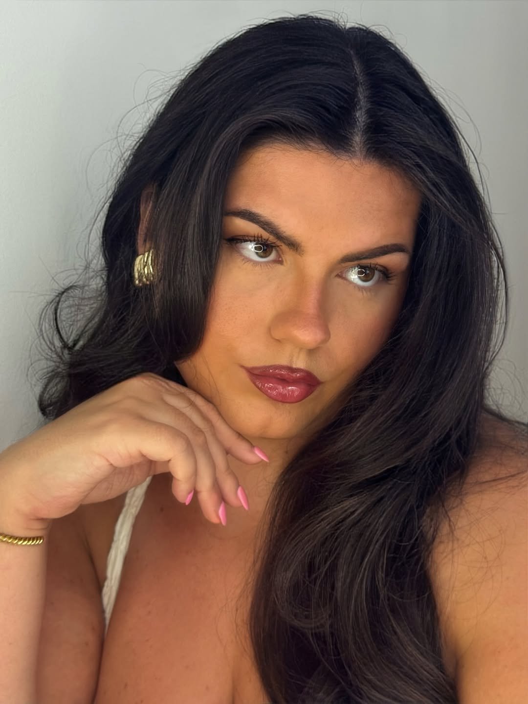 Mulher de cabelo escuro e saudável, pele impecável, olhando pensativa, com maquiagem suave e unhas rosas.