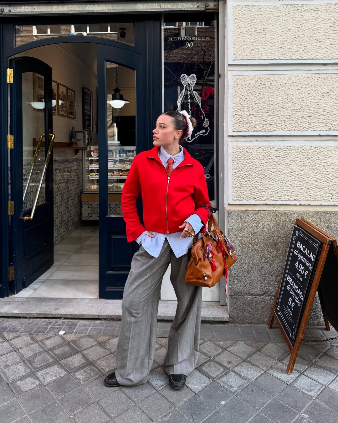 Pessoa com calça de alfaiataria cinza, camisa azul e casaco vermelho em frente a uma loja.