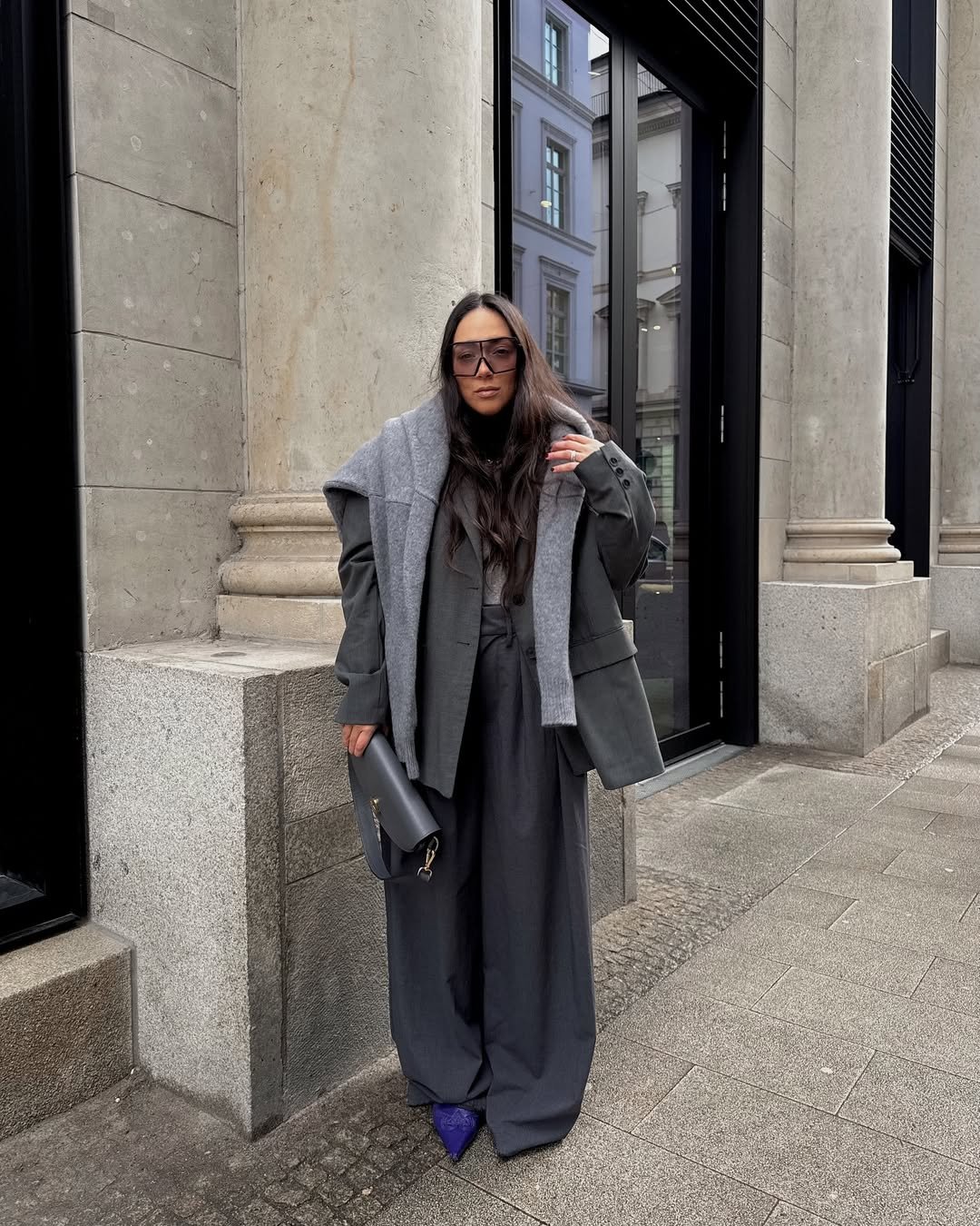 Usuária vestindo calça de alfaiataria cinza com casaco e lenço. Look elegante com bolsa cinza e sapatos roxos. Fundo urbano.