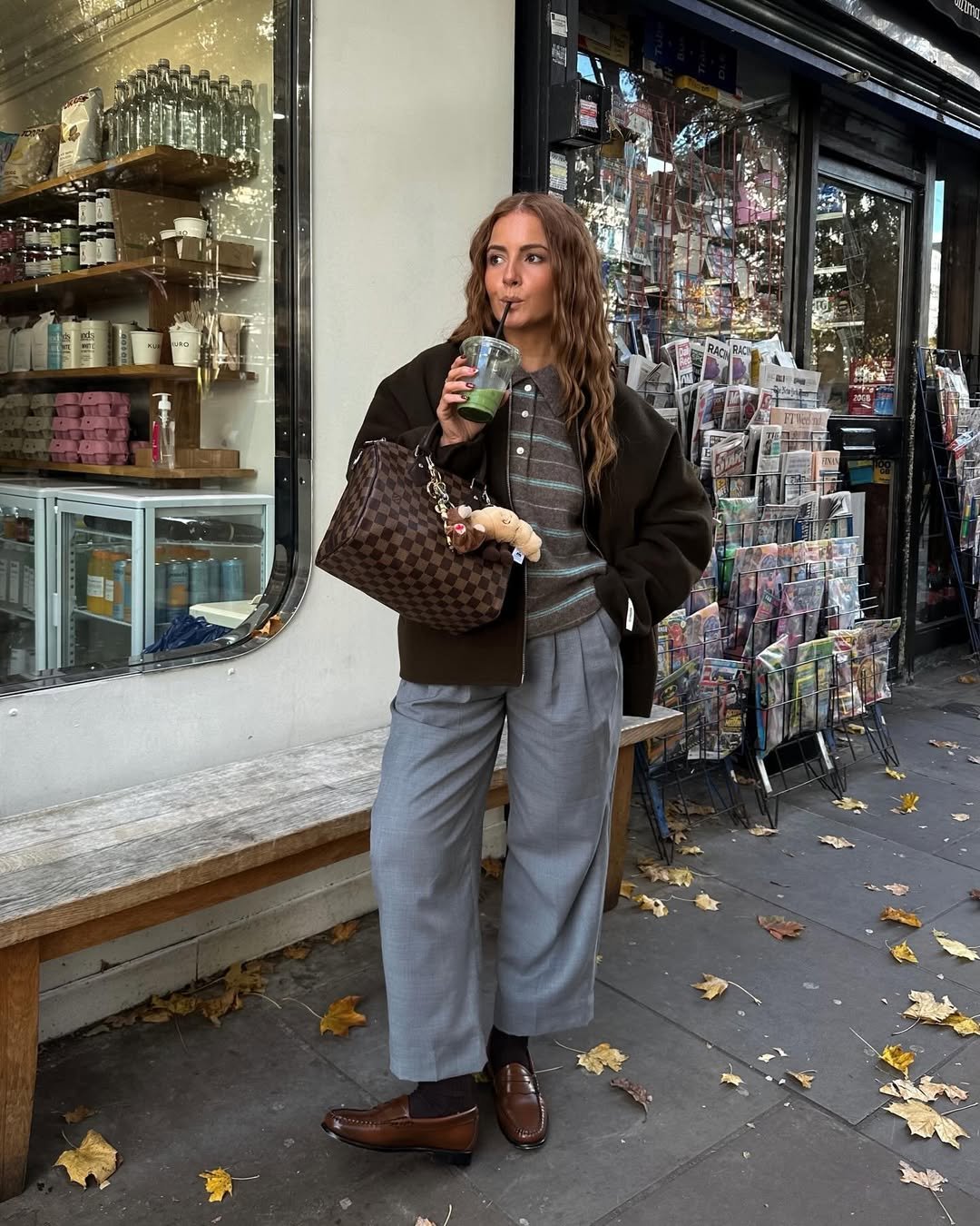 Pessoa com calça cinza, sapatos marrons e casaco escuro, segurando bebida. Loja de revistas ao fundo, mostrando como usar calça de alfaiataria cinza.