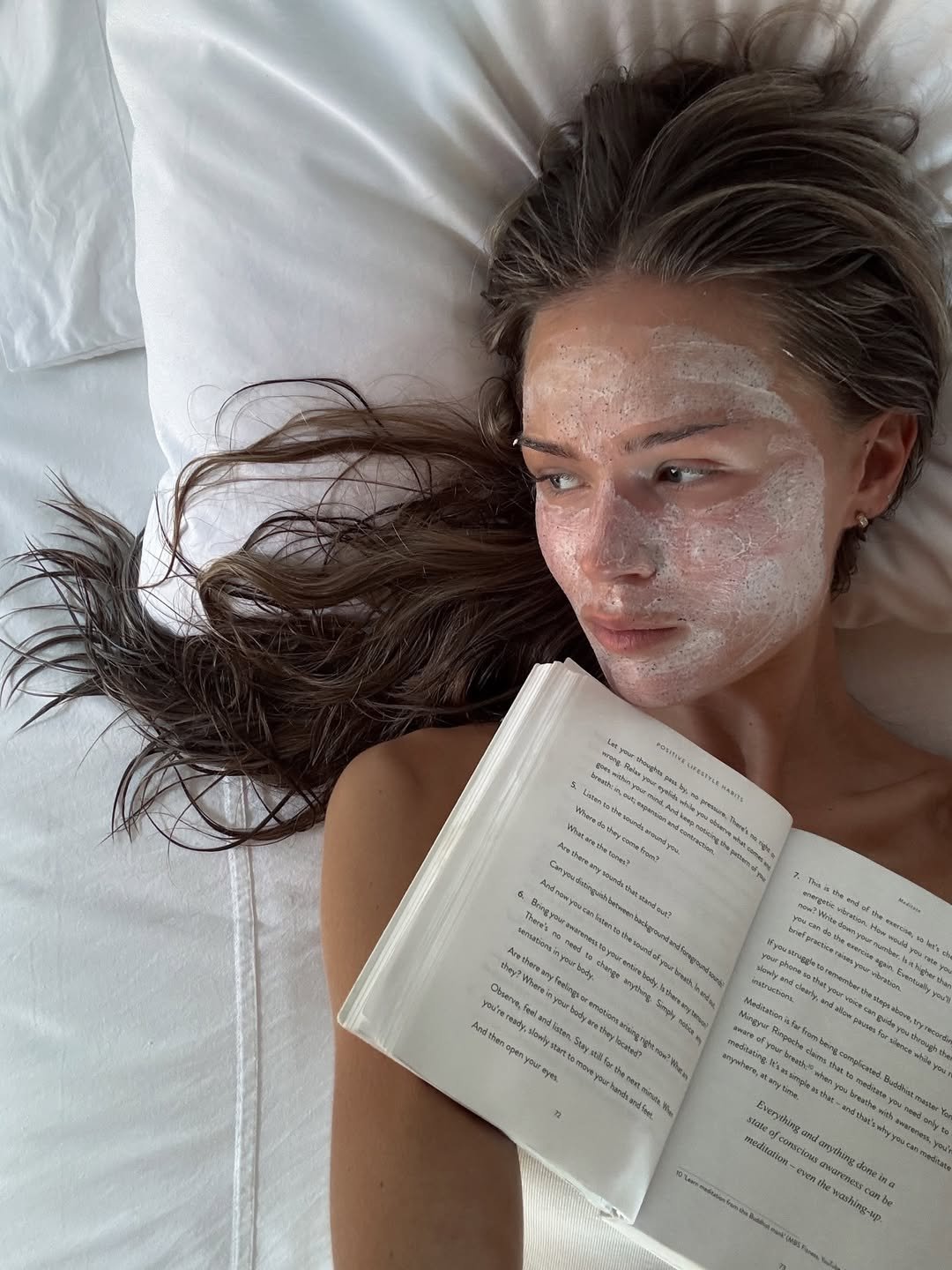 Mulher deitada na cama com máscara facial, segurando um livro. Expressão serena reflete descanso e alívio de cansaço emocional.