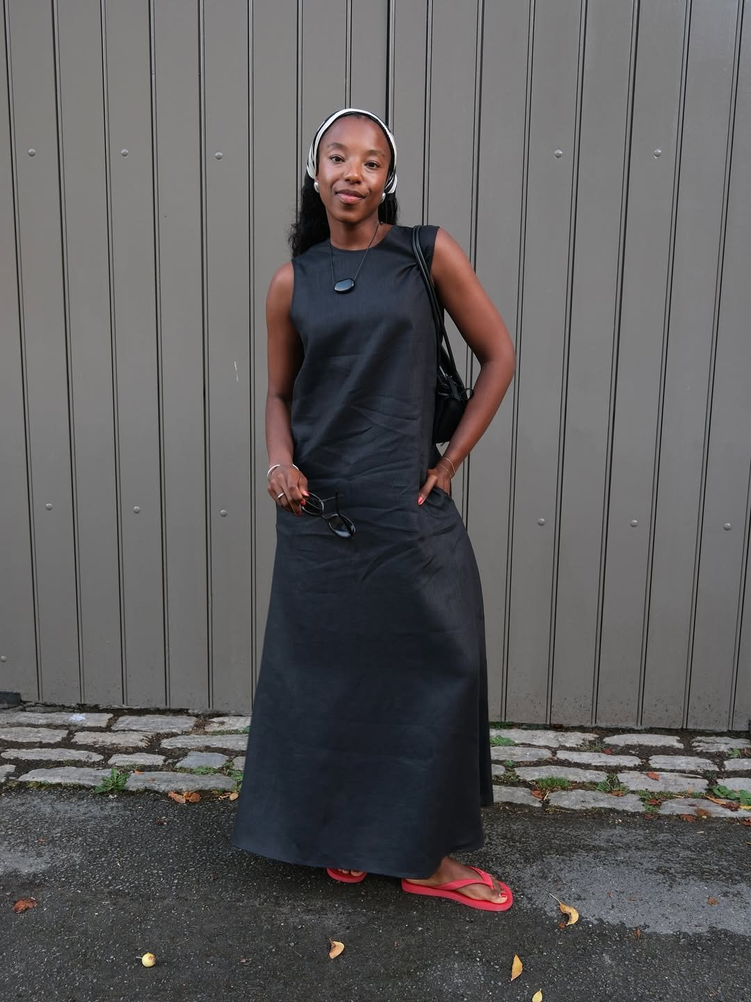Pessoa de vestido preto e chinelo vermelho posa em frente a uma parede cinza.