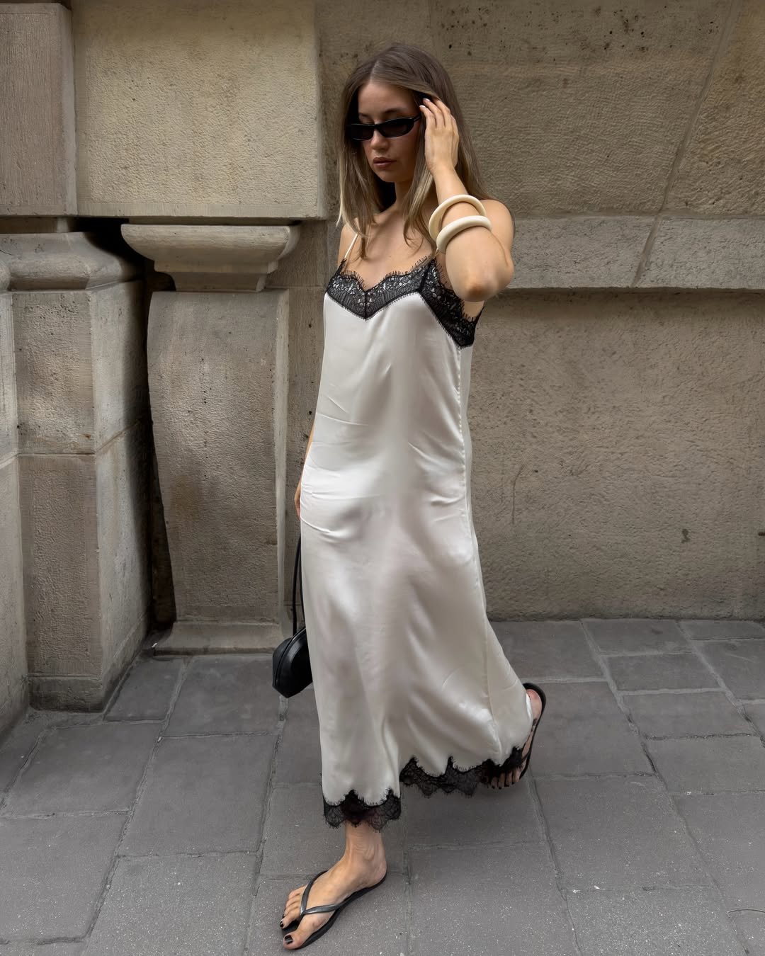 Mulher com vestido de cetim e chinelos pretos caminha em calçada de pedra, segurando uma bolsa pequena.