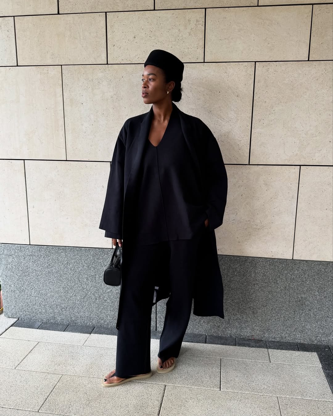 Pessoa em traje preto elegante, usando chinelos bege e segurando uma bolsa, apoiada em parede de azulejos claros.
