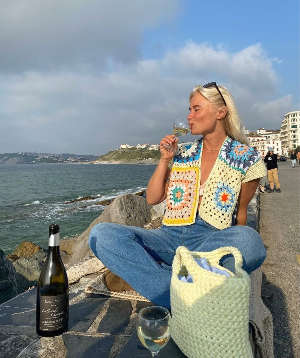 Pessoa usando um colete de crochê colorido, sentada à beira-mar, bebendo vinho.