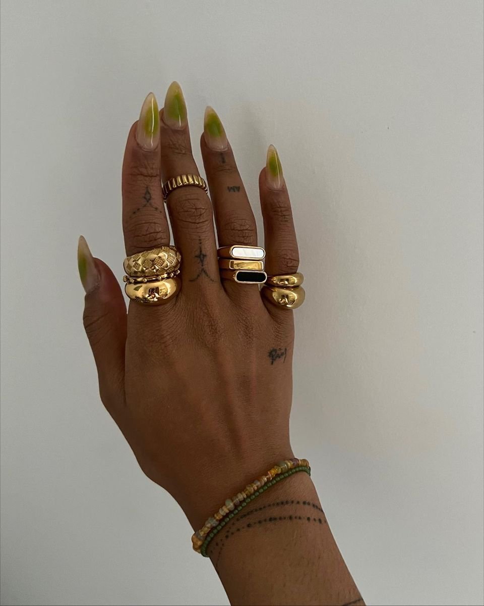 Vários anéis dourados em um mão com tatuagens e unhas compridas. Como combinar anéis