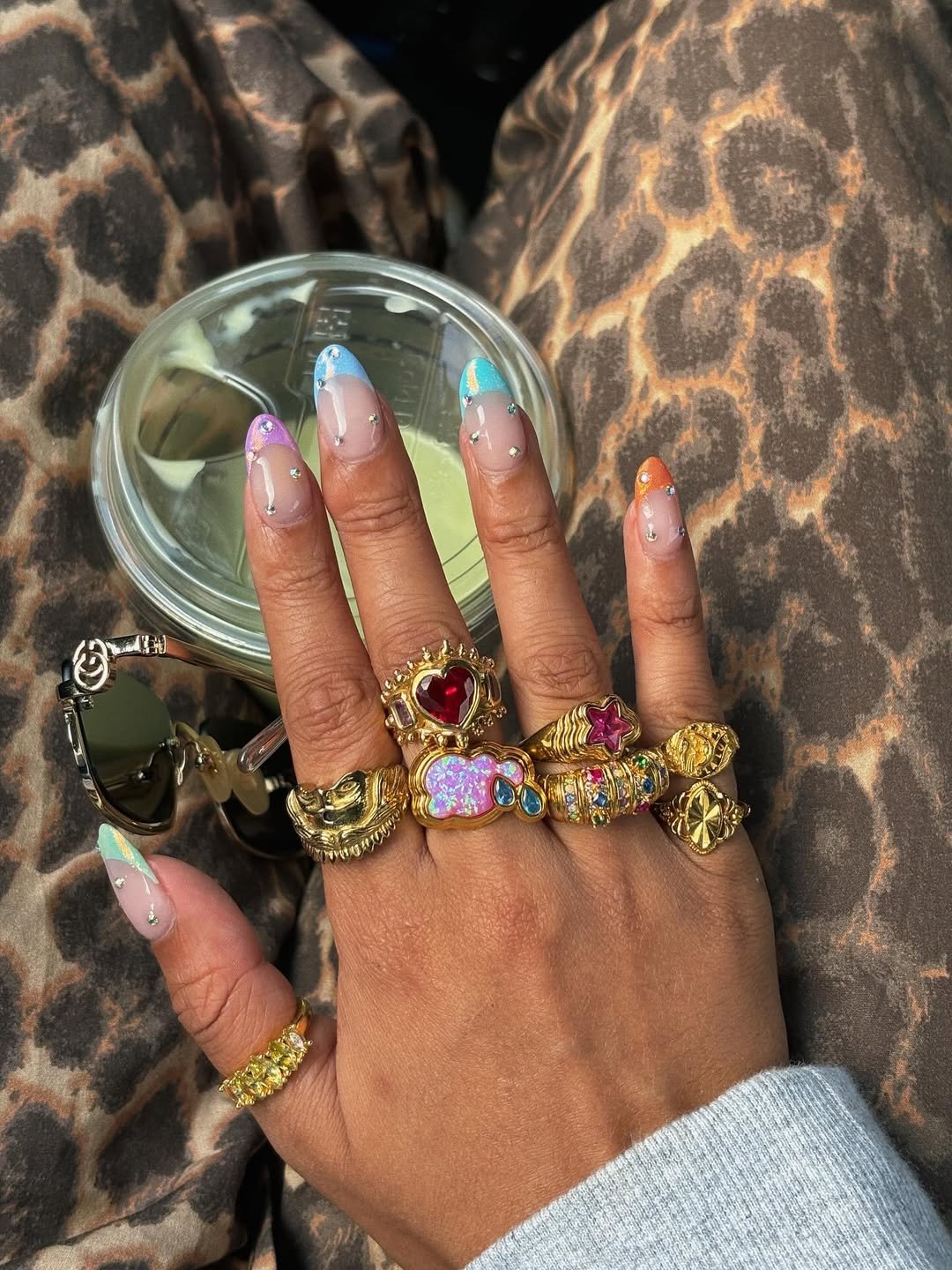 Unhas coloridas e mãos repletas de anéis dourados e rebuscados sobre calças estampadas de leopardo.