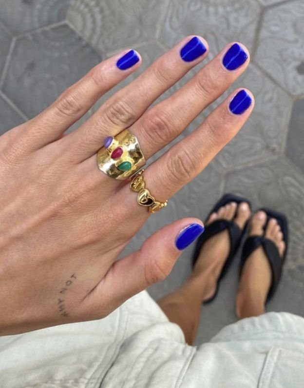Unhas das mãos pintadas de azul, com anéis dourados; ao fundo, pés em sandálias.