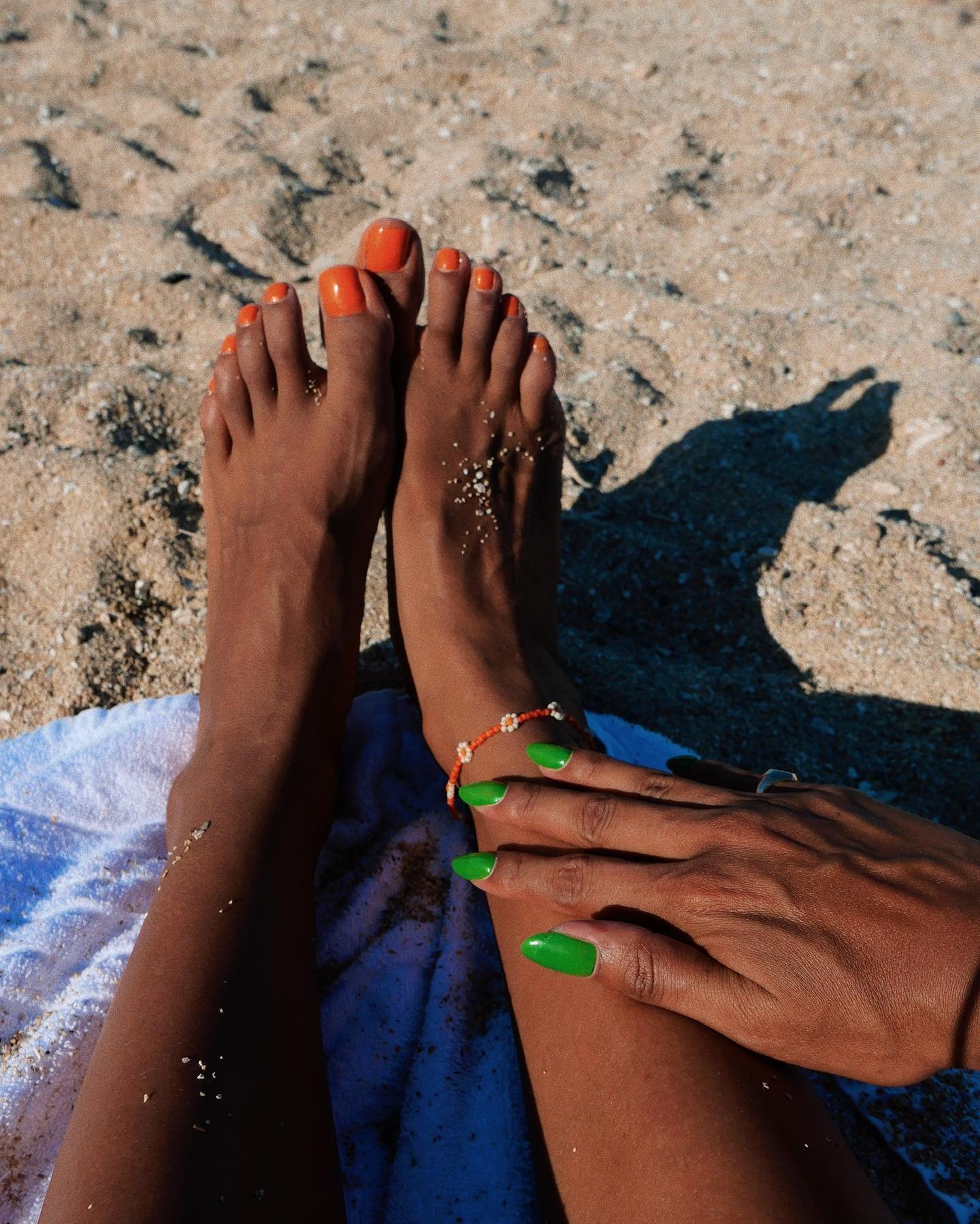 Unhas do pé pintadas de laranja na areia; mãos com unhas verdes tocam o tornozelo.