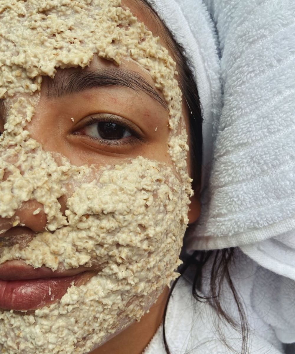 Rosto coberto por máscara esfoliante de aveia, focado na área dos olhos, enquanto esfoliar o rosto com suavidade.