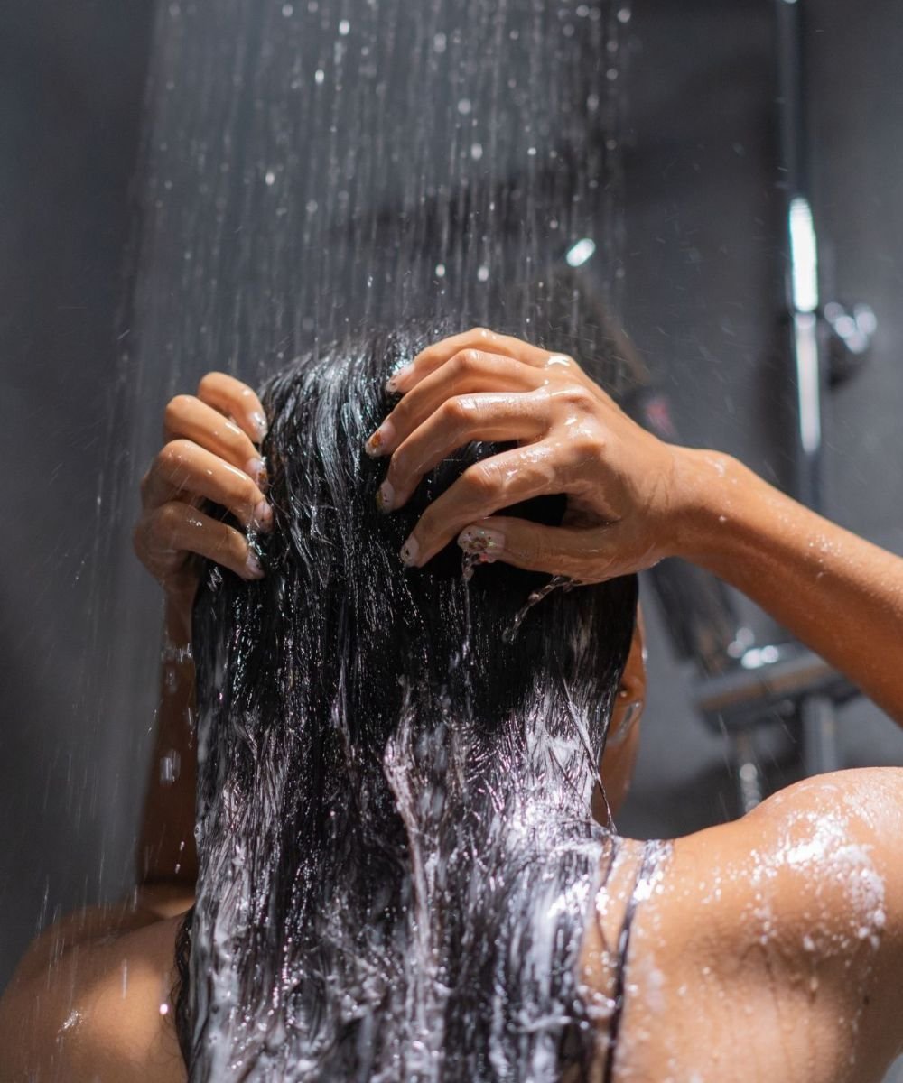 Pessoa lavando os cabelos sob o chuveiro, aplicando shampoo caseiro com as mãos molhadas.