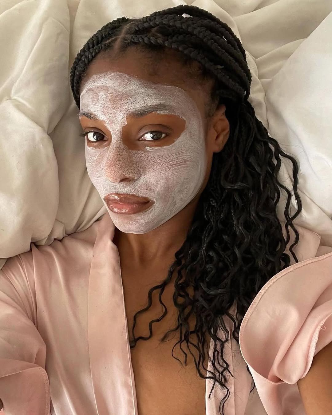 Pessoa deitada com máscara facial branca, vestindo roupão rosa claro e cabelo trançado sobre travesseiros.