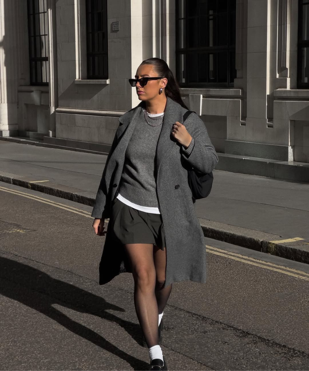 Pessoa caminhando na rua com casaco cinza sobre vestido, exibindo estilo casual sofisticado.