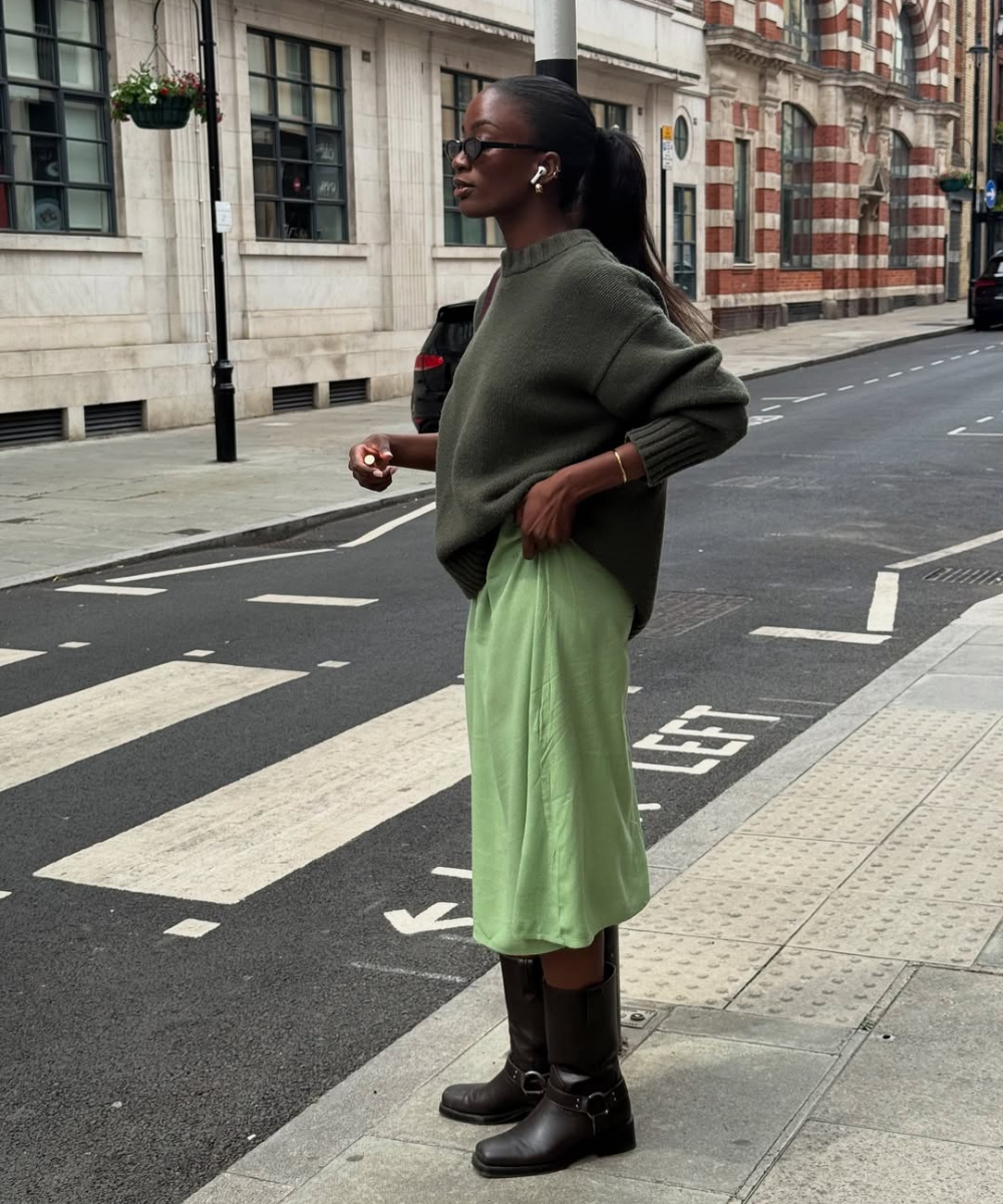 Pessoa com óculos escuros e cabelo preso usa suéter verde e saia verde-clara, parada na calçada urbana.