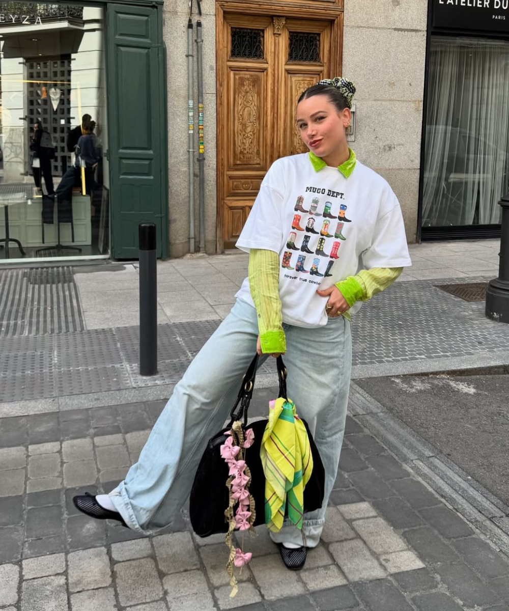 Jovem com camisa gráfica, sobre camisa verde, calça jeans larga e bolsa com lenços amarrados.