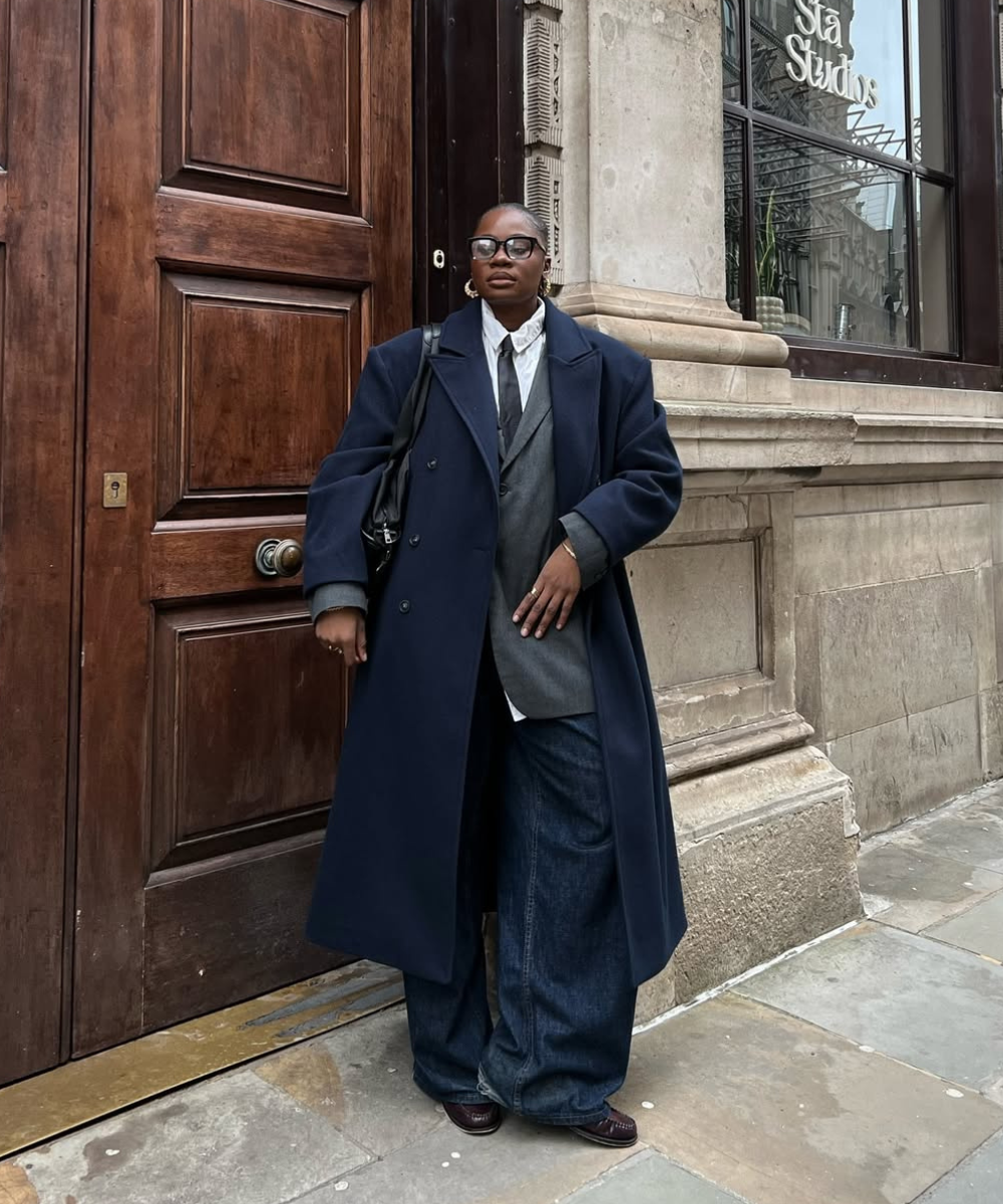 Pessoa de pé, vestindo casaco azul sobre terno cinza, mostrando como usar sobreposições de roupas estilosas.