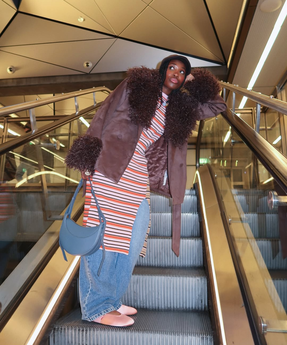 Pessoa vestida com casaco marrom e vestido listrado, subindo escada rolante, exibindo sobreposições estilizadas.