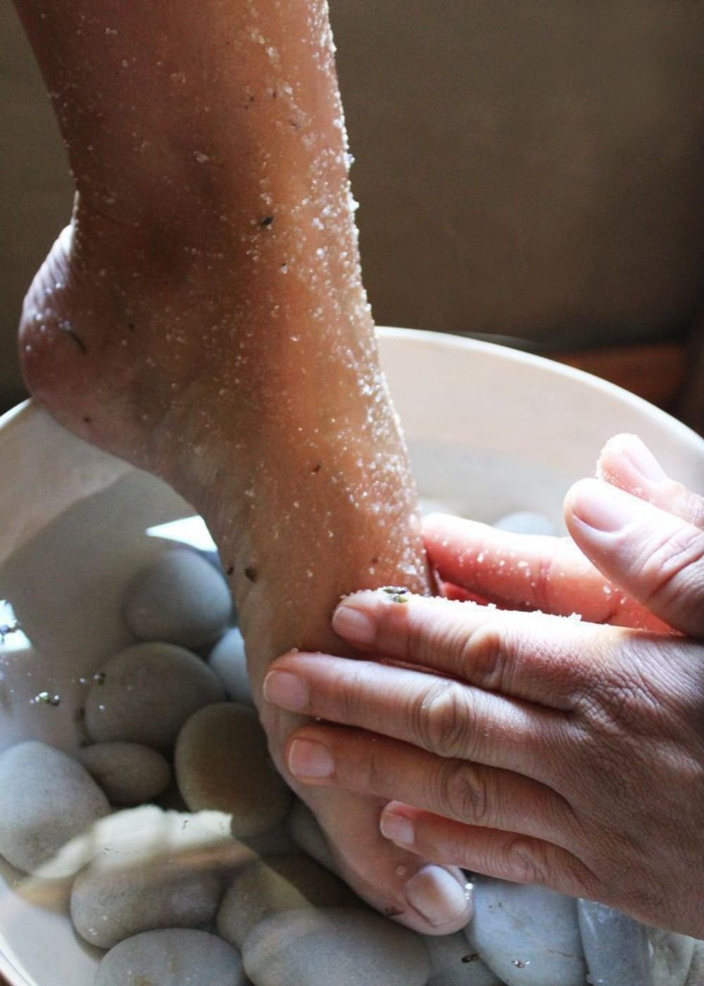 Pé sendo esfoliado com sal grosso durante um escalda-pés em uma bacia com pedras lisas.