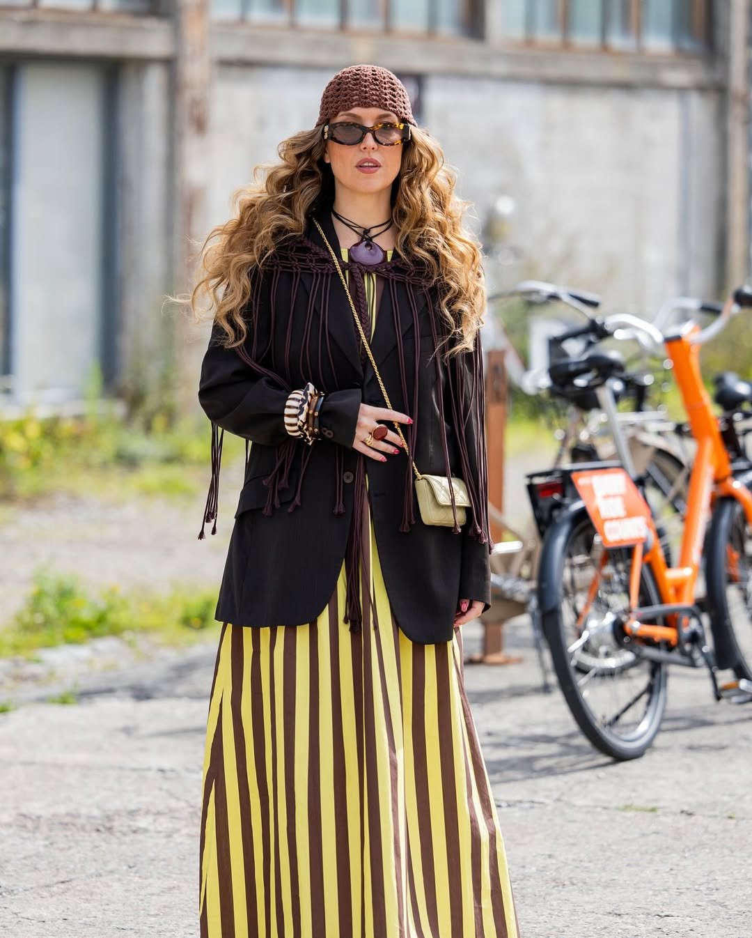 Mulher elegante com touca de crochê marrom, usando óculos escuros, casaco escuro e saia listrada ao fundo de bicicletas.