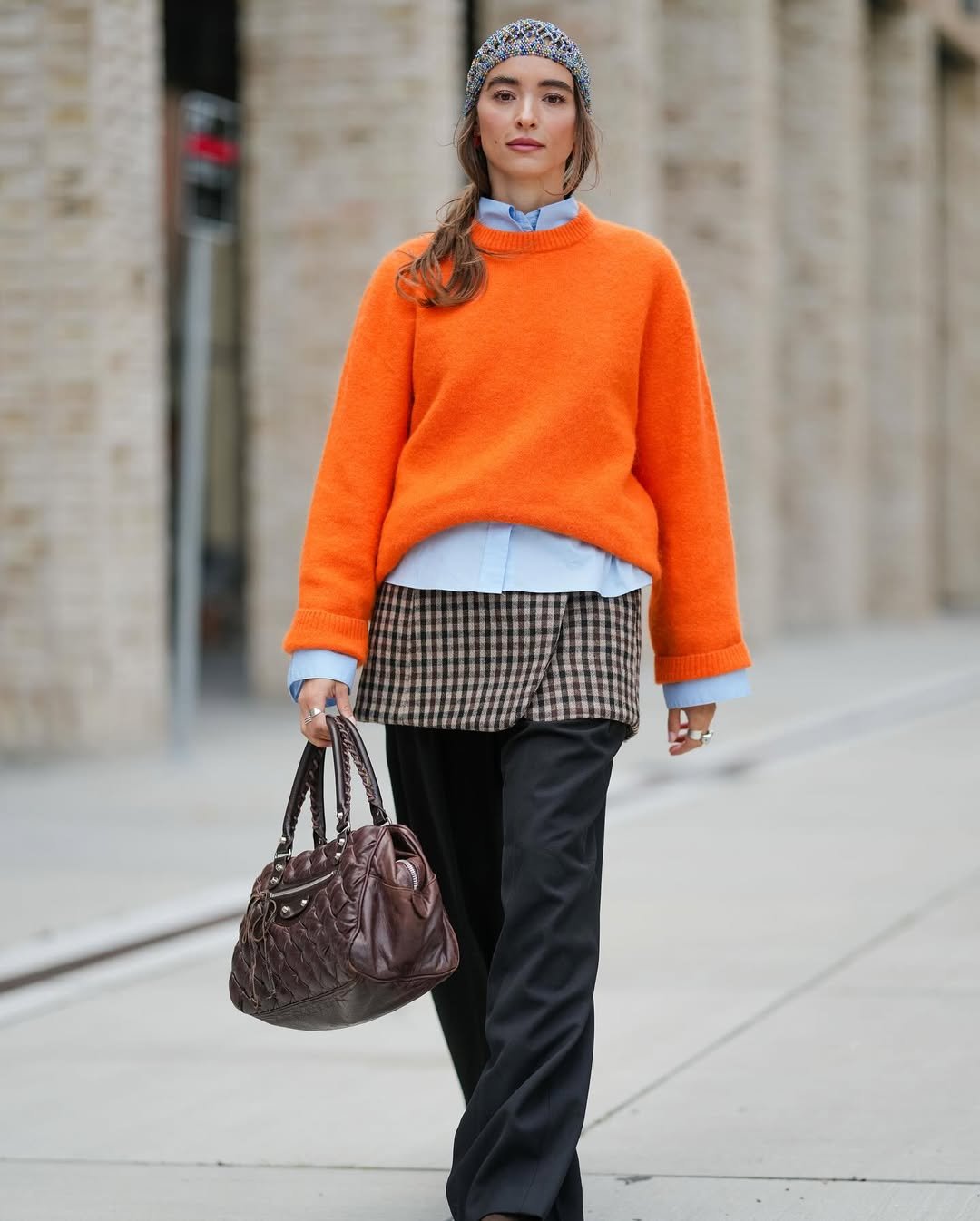 Mulher vestida com touca de crochê, suéter laranja, camisa azul, saia xadrez, calça preta e bolsa marrom.