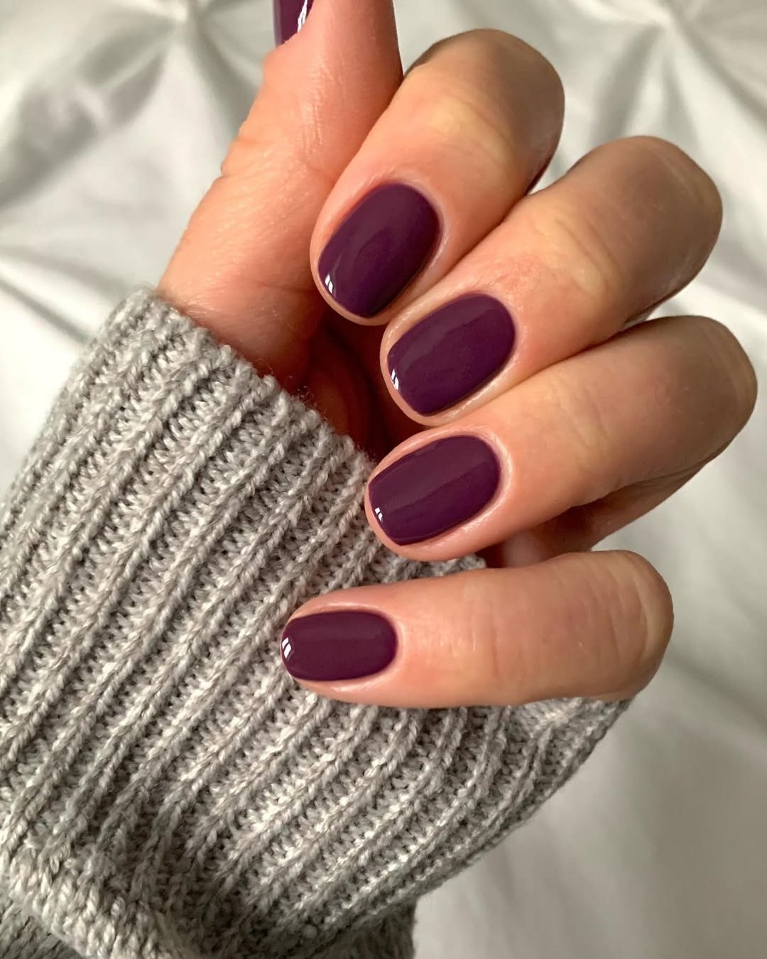 Unhas curtas esmaltadas em tom de roxo vibrante, tendência de cores de esmalte 2025, destacam-se sobre suéter cinza.