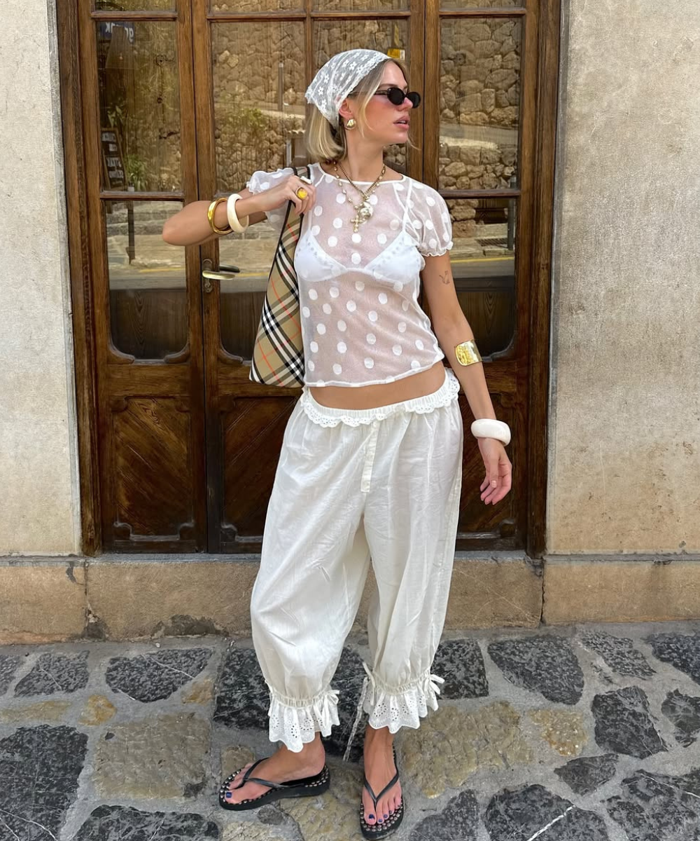 Pessoa usando roupas brancas transparentes, estilo verão, em frente a uma porta de madeira. Elementos estilosos incluem acessórios modernos.