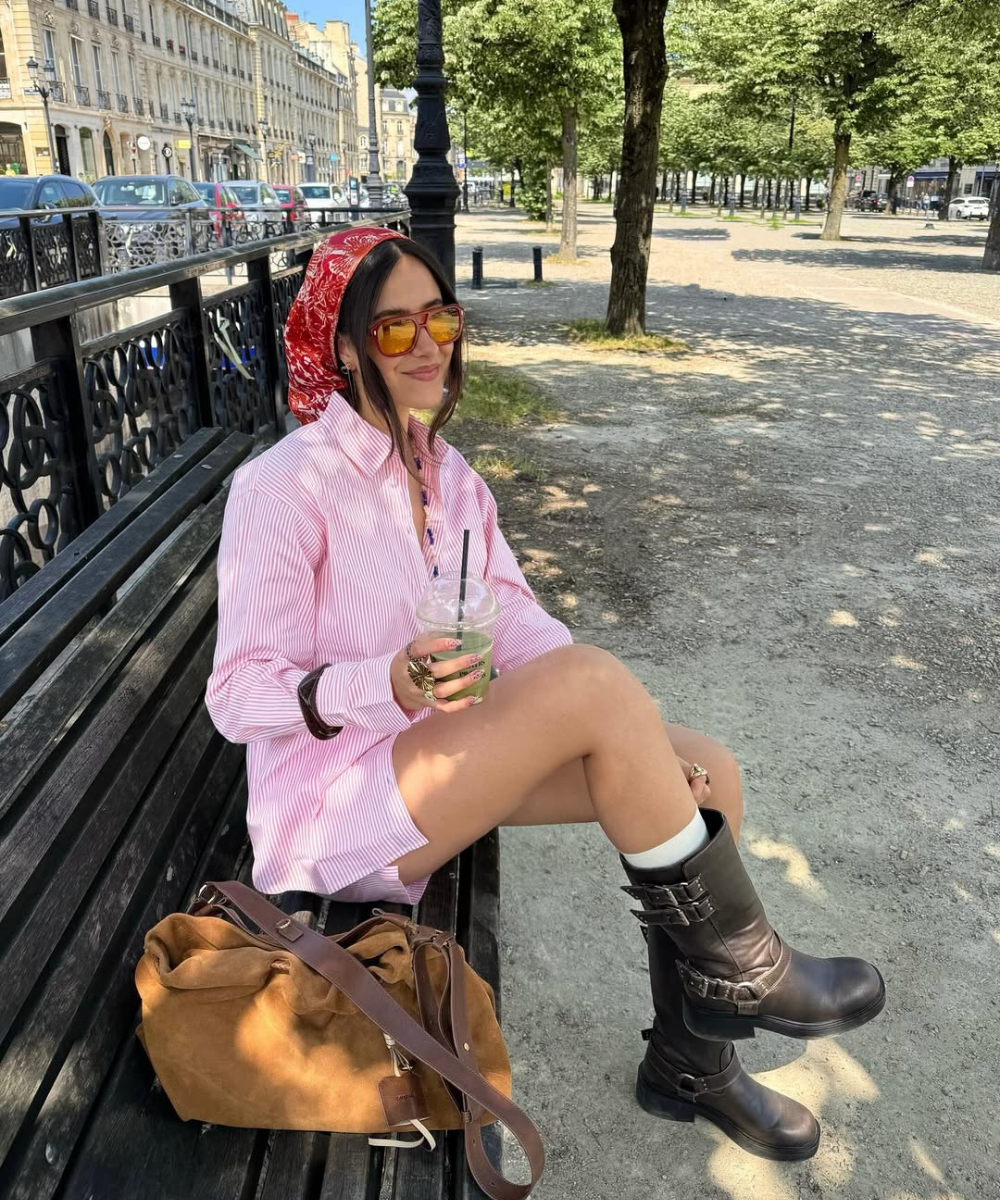 A mulher está sentada em um banco, usando botas e camiseta listrada, com cores do verão 2026.