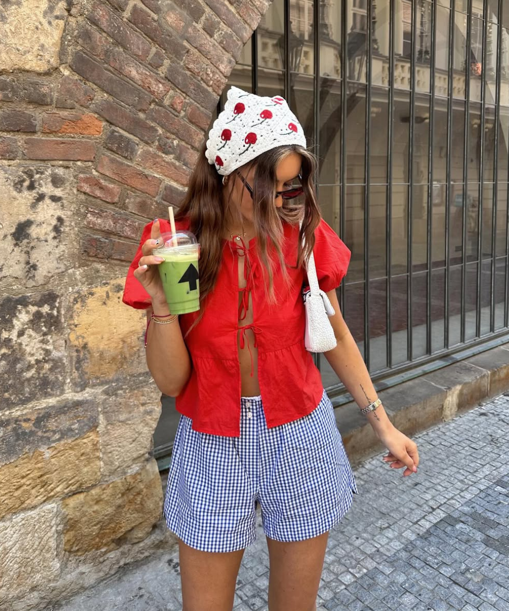 Mulher vestindo bata vermelha, short quadriculado azul, bandana de morangos e bolsa de ombro branca.