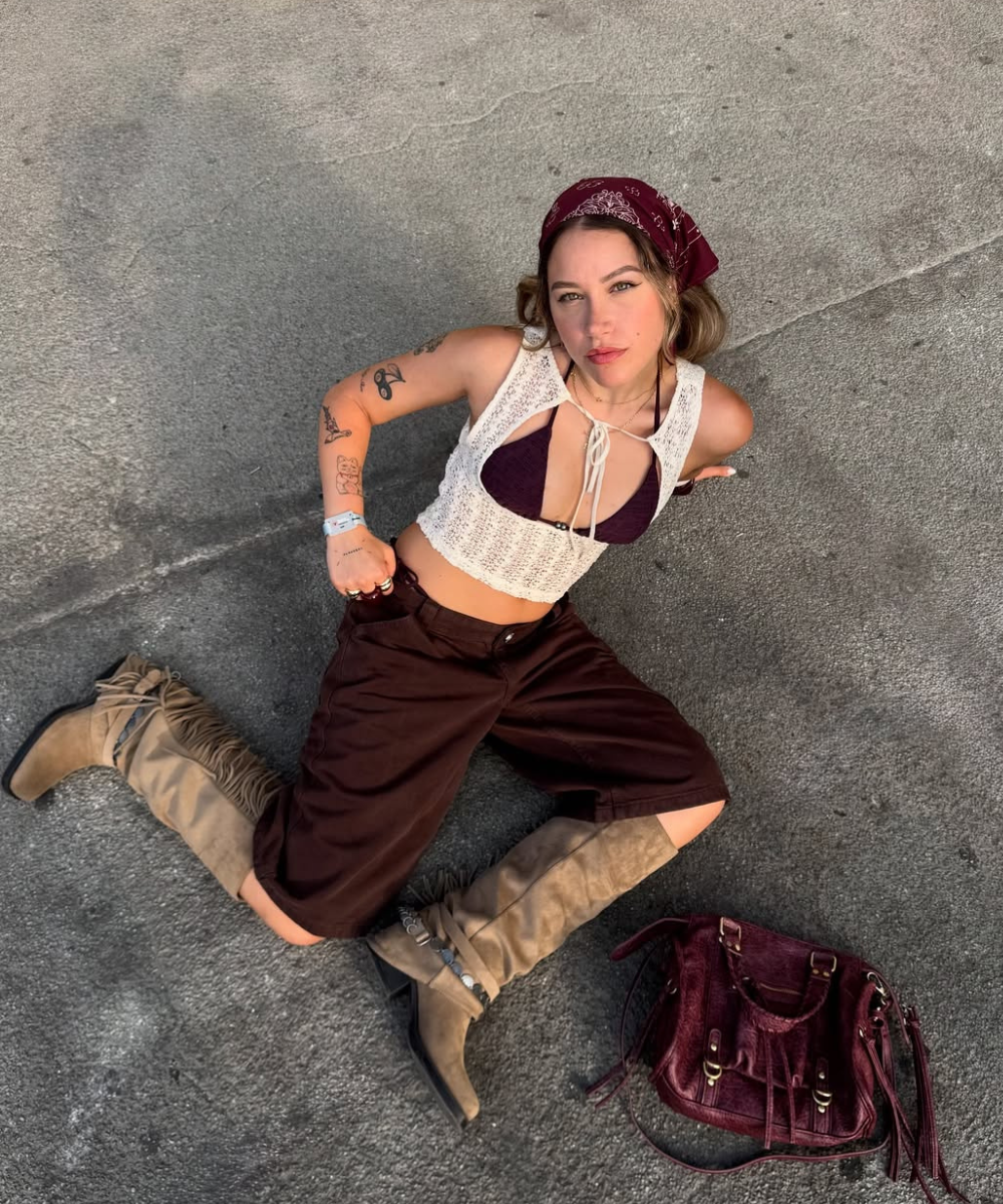Mulher sentada no chão, usando roupas marrom e creme, bota bege e lenço vermelho. Bolsa vinho ao lado.
