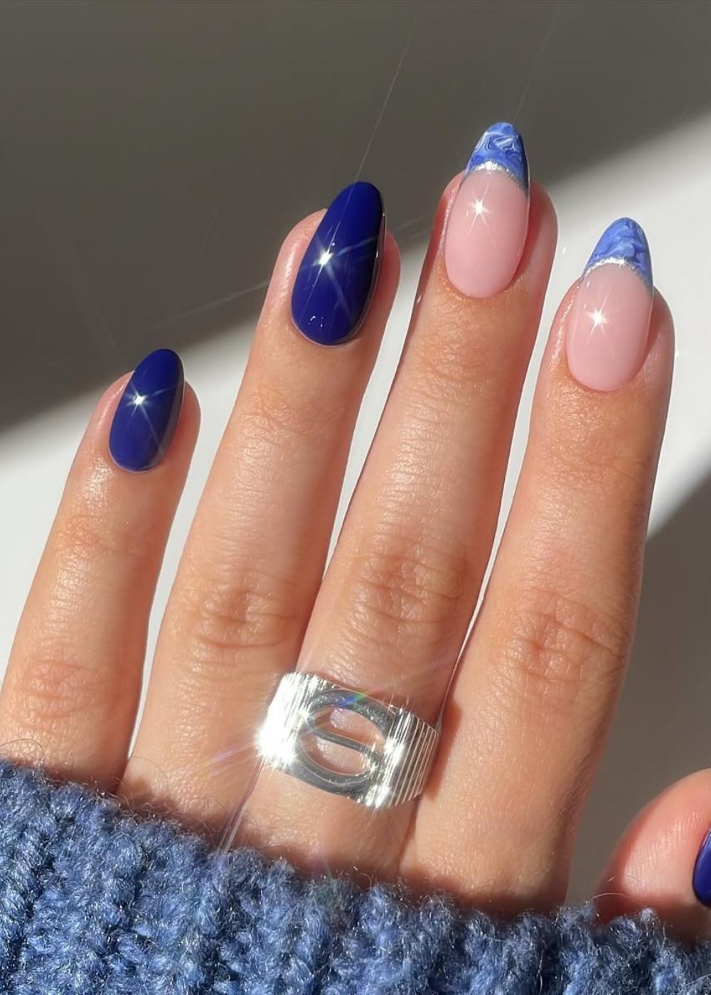 Unhas esmaltadas em azul cobalto e nude com detalhes marmorizados, destacadas por um anel prateado e uma blusa azul.
