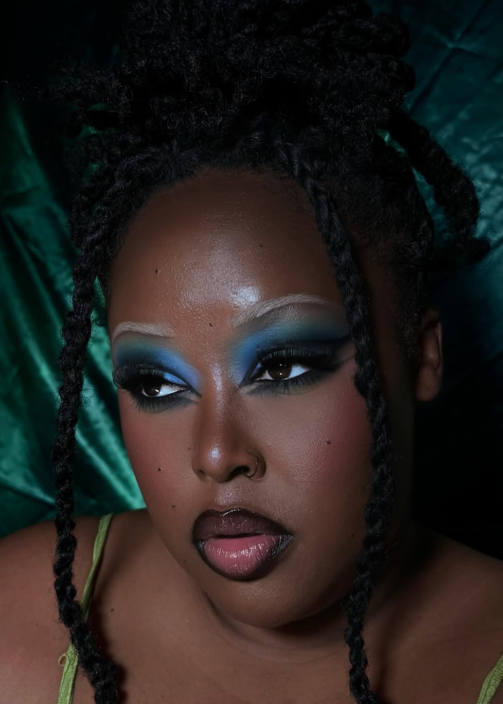 Maquiagem artística com sombra azul cobalto, destacando os olhos. Cabelo trançado, fundo verde escuro.