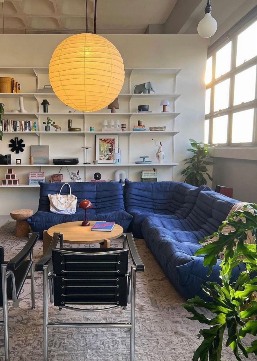 Sala de estar com sofá azul cobalto, mesa de centro redonda, luminária de papel e estante cheia de objetos e livros ao fundo.