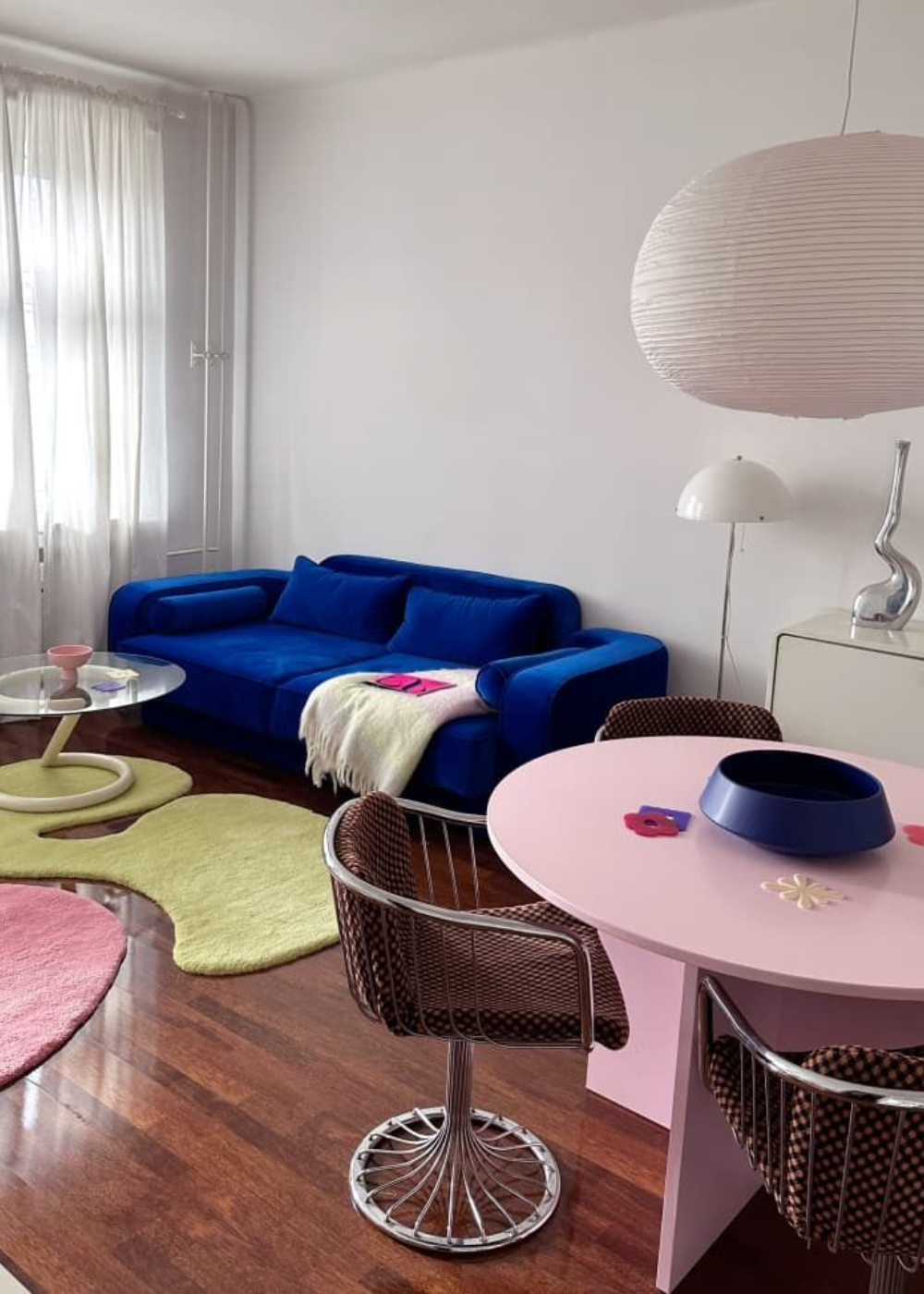 Sofá azul cobalto em sala moderna com mesa rosa, tapetes coloridos e luminária branca.