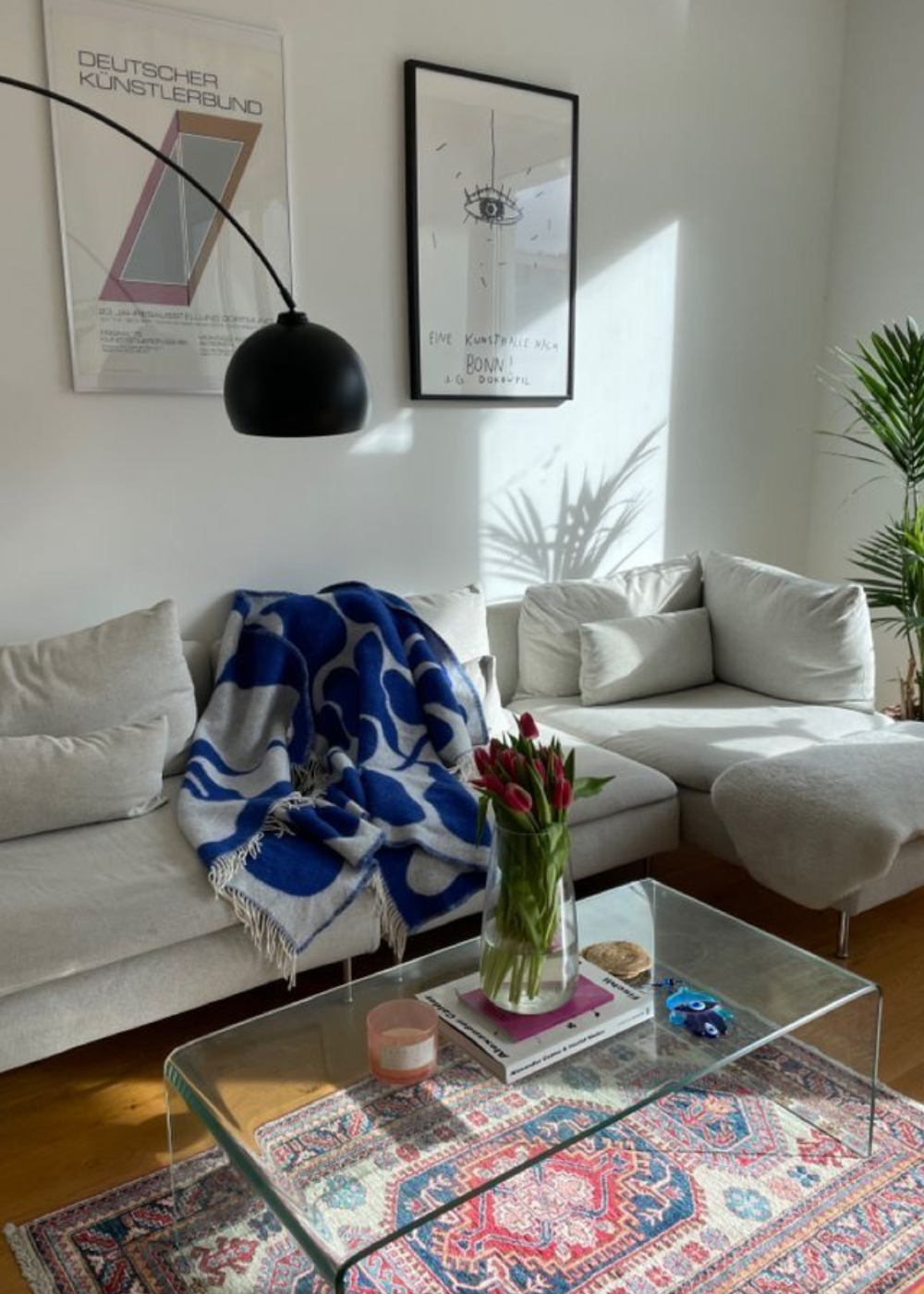 Sala de estar com sofá cinza, cobertor azul cobalto, mesa de vidro com tulipas e pôsteres na parede.