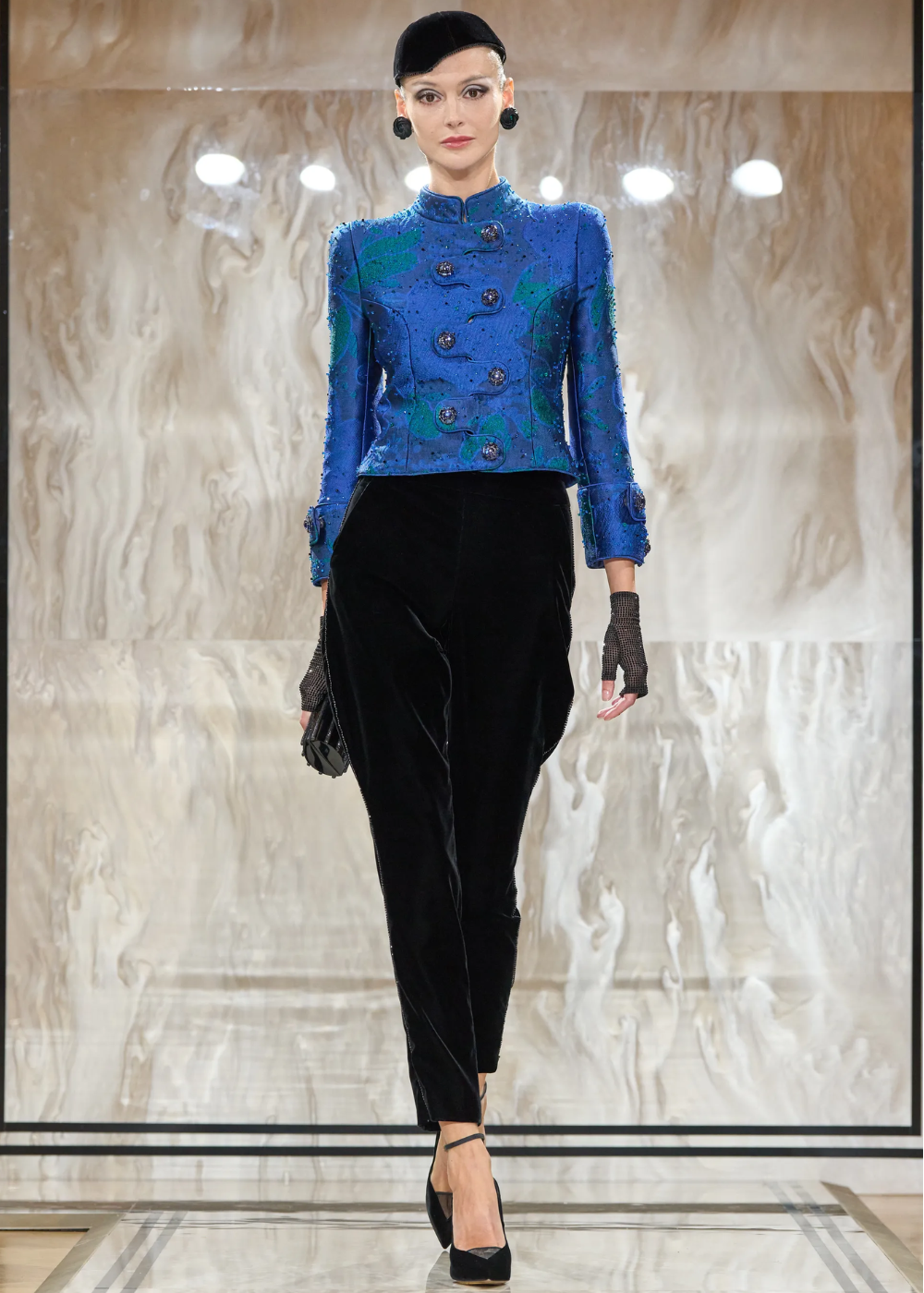 Modelo exibe traje elegante com casaco azul cobalto e calça preta em passarela de mármore.