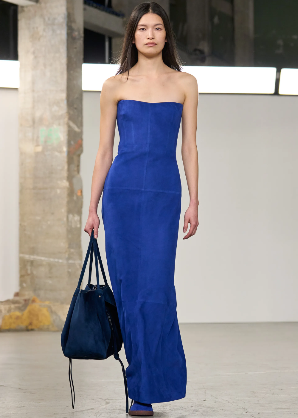 Modelo usando vestido longo azul cobalto, segurando bolsa combinando em ambiente industrial.