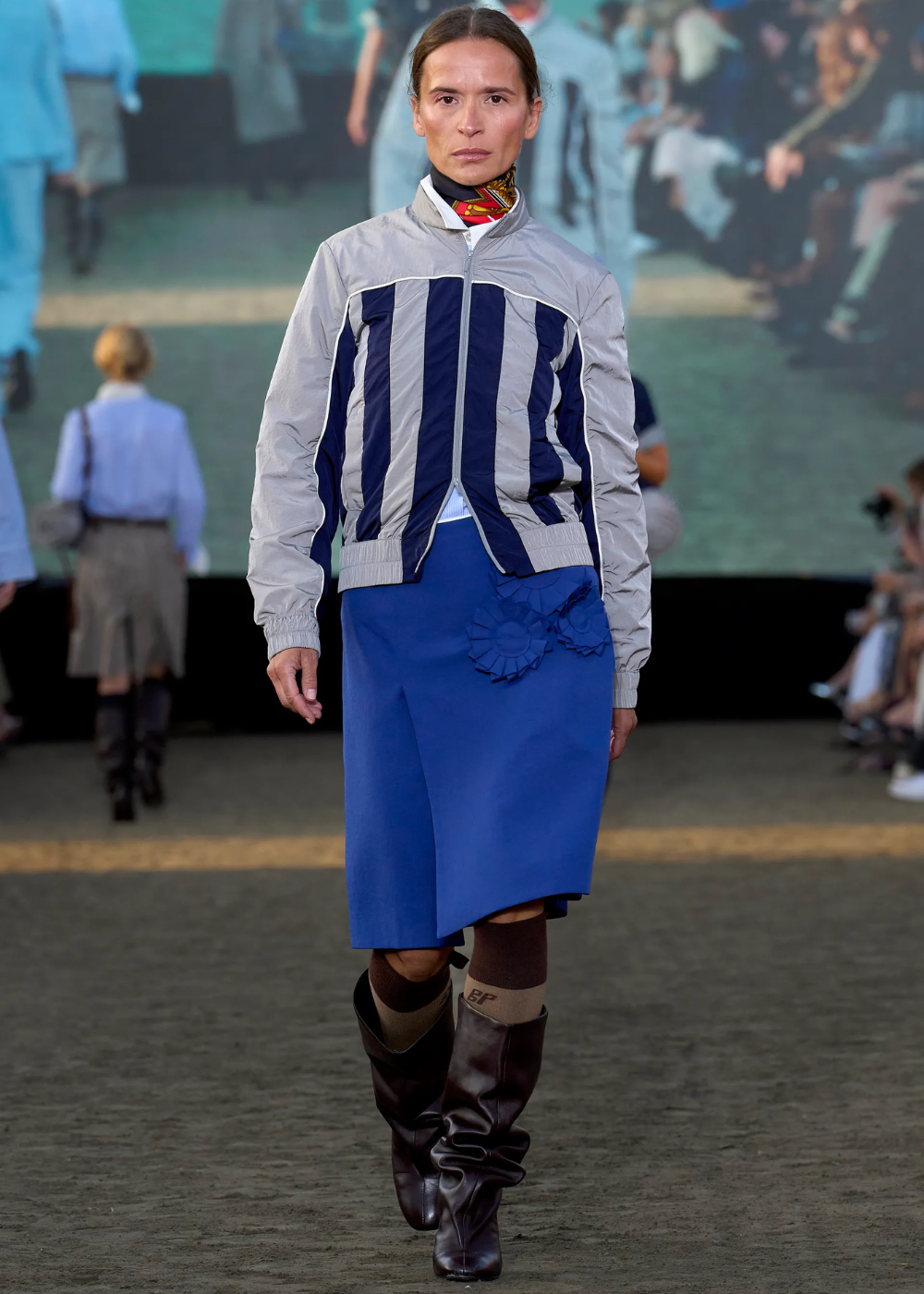 Modelo com saia azul cobalto e jaqueta listrada, desfilando com botas escuras em uma passarela.