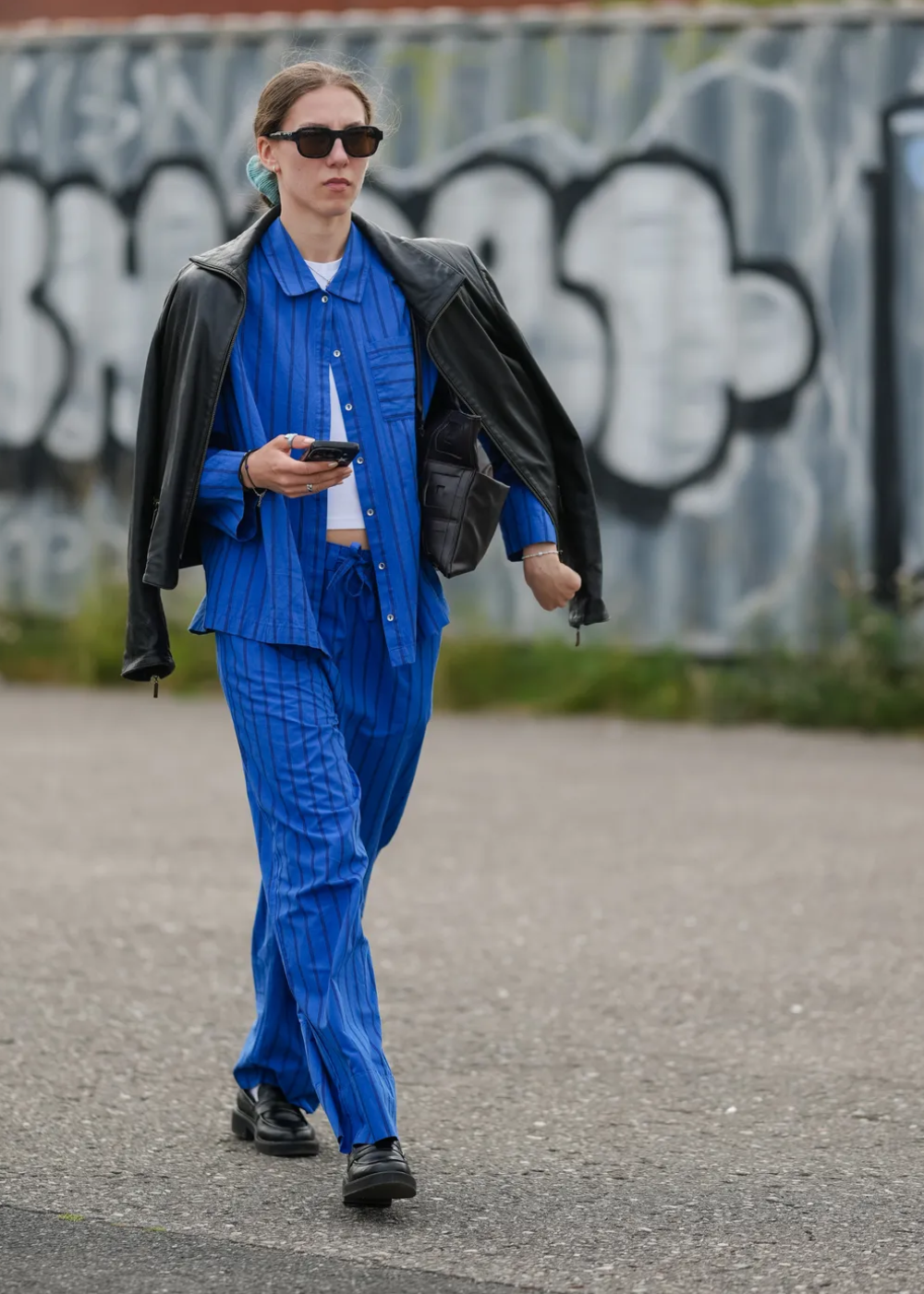 Pessoa usando conjunto azul cobalto com blazer preto nos ombros, segurando celular, em frente a mural grafitado.