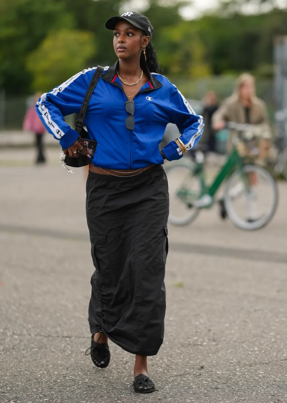 Pessoa caminhando com um casaco azul cobalto e boné preto, segurando um celular. Ao fundo, uma bicicleta verde.