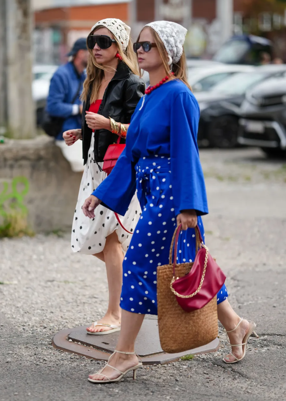 Duas mulheres caminhando, uma delas veste azul cobalto com estampa de poás, segurando bolsa vermelha. A outra usa preto e branco.